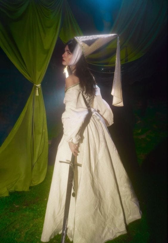 White medieval dress with gold trim, matching hat, holding a sword against green fabric.