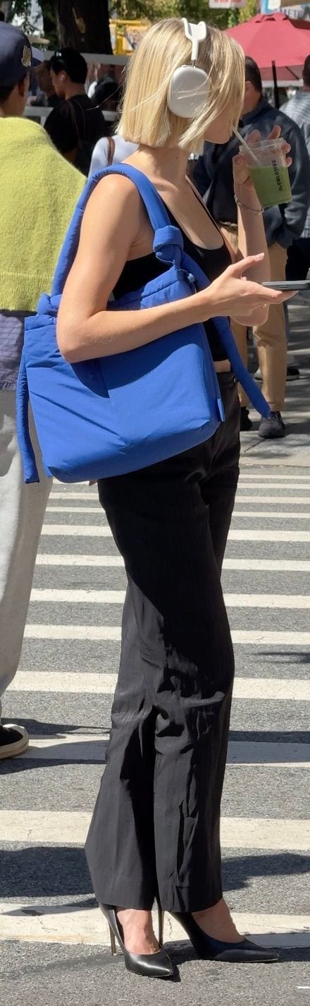 Blonde woman in black top, black pants, black heels, blue padded bag, white headphones.