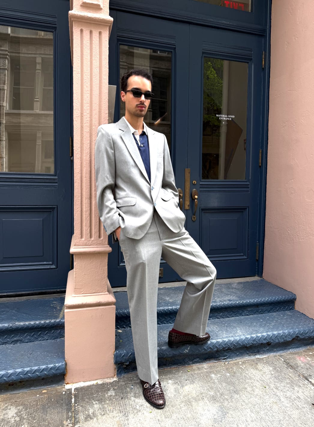 Light grey suit with a navy polo shirt and black sunglasses, outside a blue door.