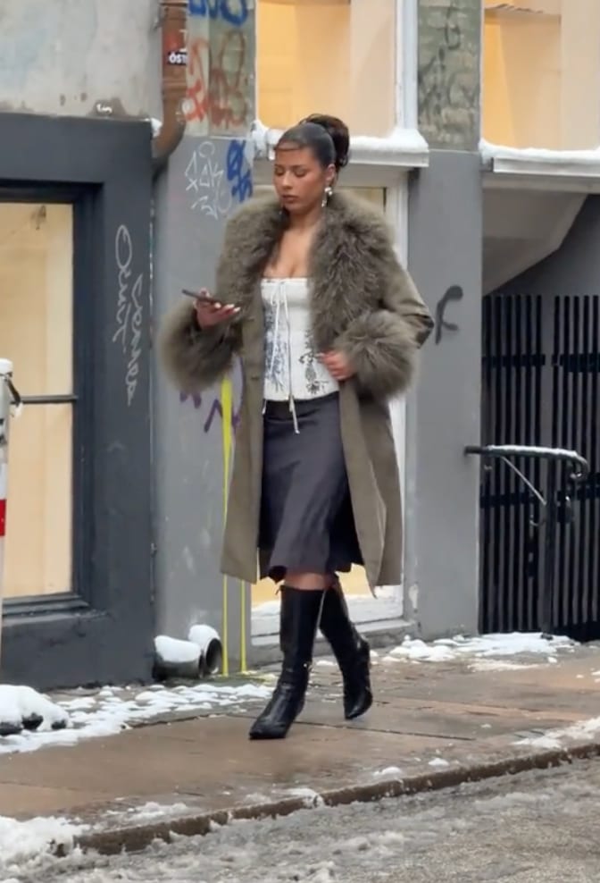 Green fur-trimmed coat over a white corset and dark skirt, with black knee-high boots.