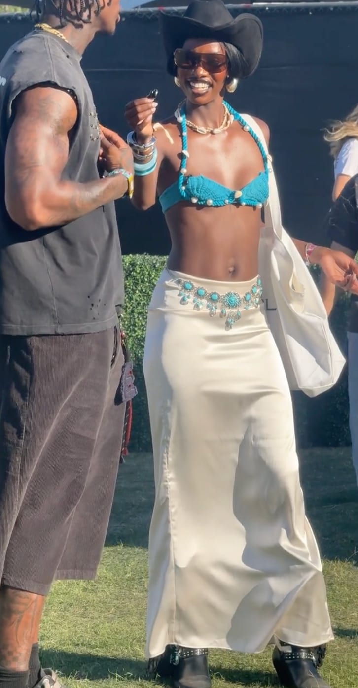Black cowboy hat, teal crochet top, off-white satin skirt with turquoise belt, and black boots.