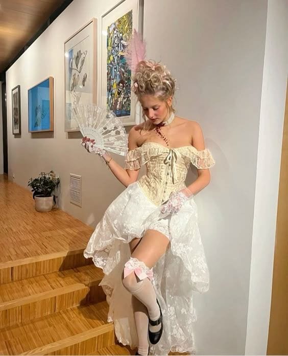 Cream off-shoulder corset, white lace skirt, sheer stockings, and a feathered fan.
