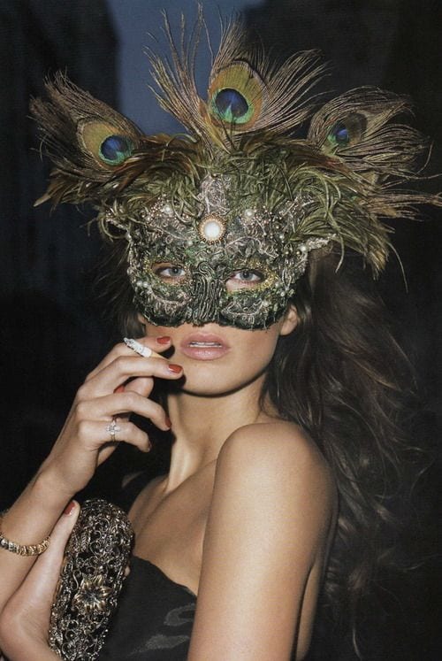 Woman in peacock feather mask, strapless top, holding a cigarette and ornate clutch.