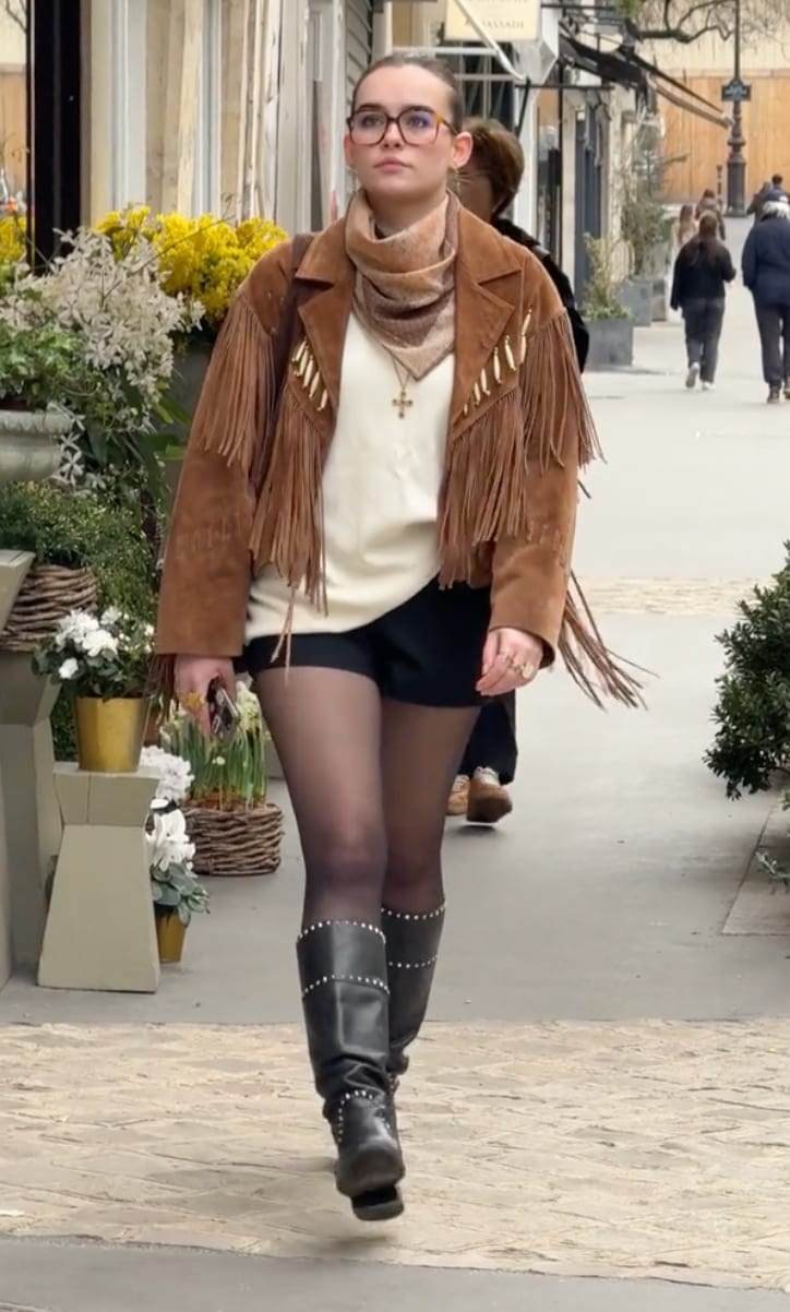 Brown suede fringe jacket, white top, black shorts, black tights, and studded boots.
