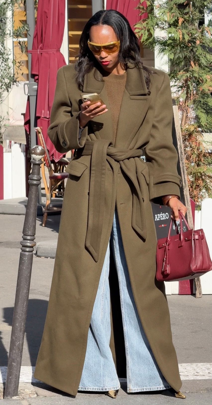 Khaki longline trench coat with wide-leg jeans, holding a burgundy handbag and phone.