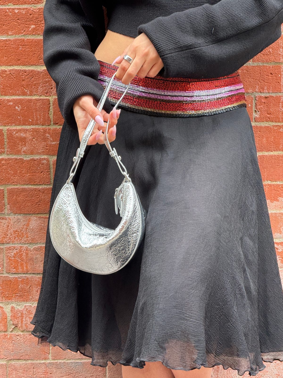 Black ribbed crop top and sheer black skirt with a silver crescent bag against a brick wall.