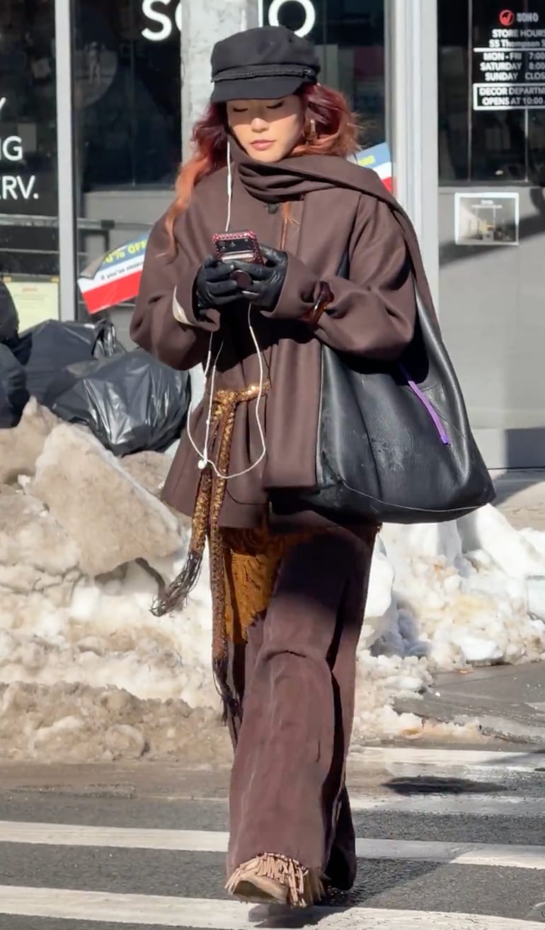 Brown coat with golden patterned scarf, black hat, gloves, and handbag on a snowy street.