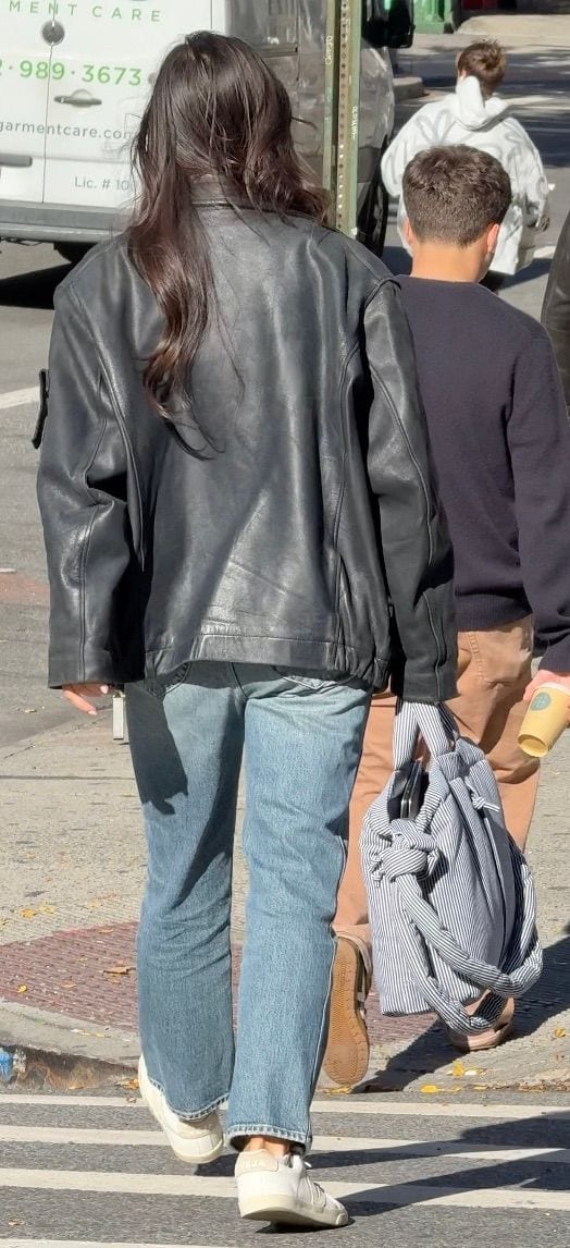 Black leather jacket with blue jeans and white sneakers walking down a city street.