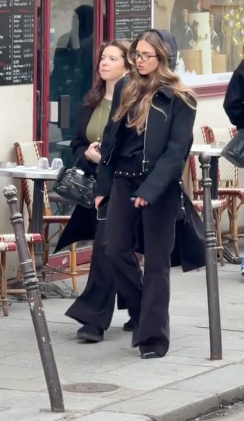 Black zip-up jacket over dark flared pants, glasses, and a black handbag on a street.