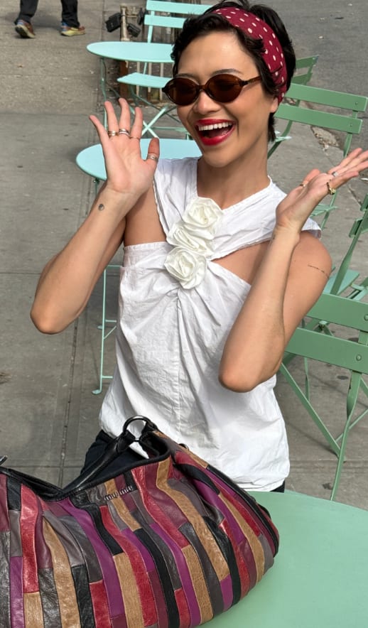 White asymmetrical top with floral detail, red polka dot headband, brown sunglasses, and colorful patchwork bag.
