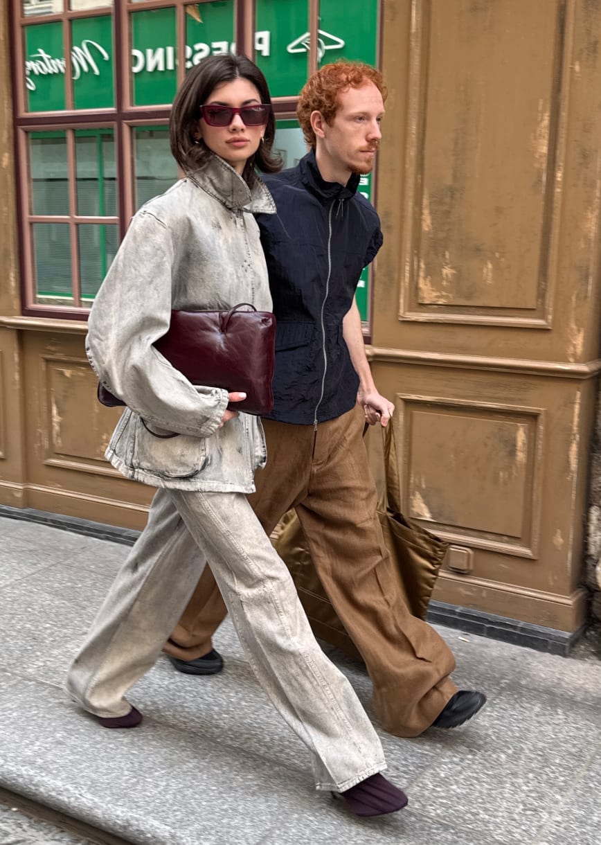 Washed grey denim jacket and jeans, burgundy clutch, dark sunglasses; man in black top, brown pants.