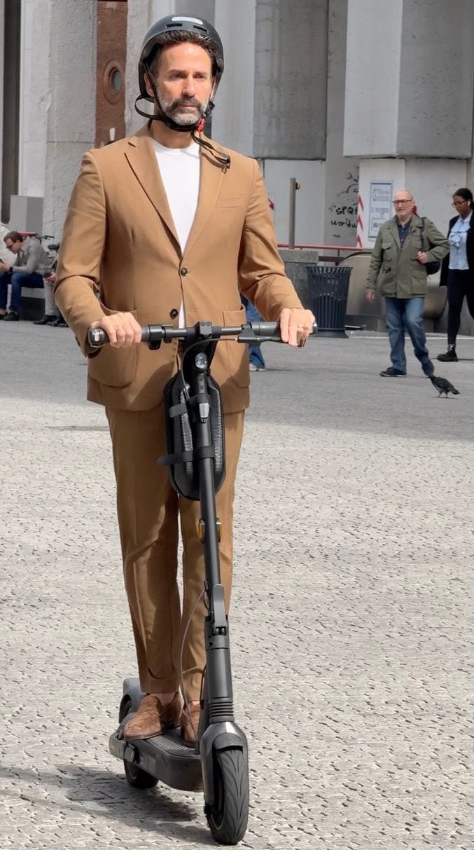 Brown suit and white t-shirt with a black helmet, riding a black electric scooter.