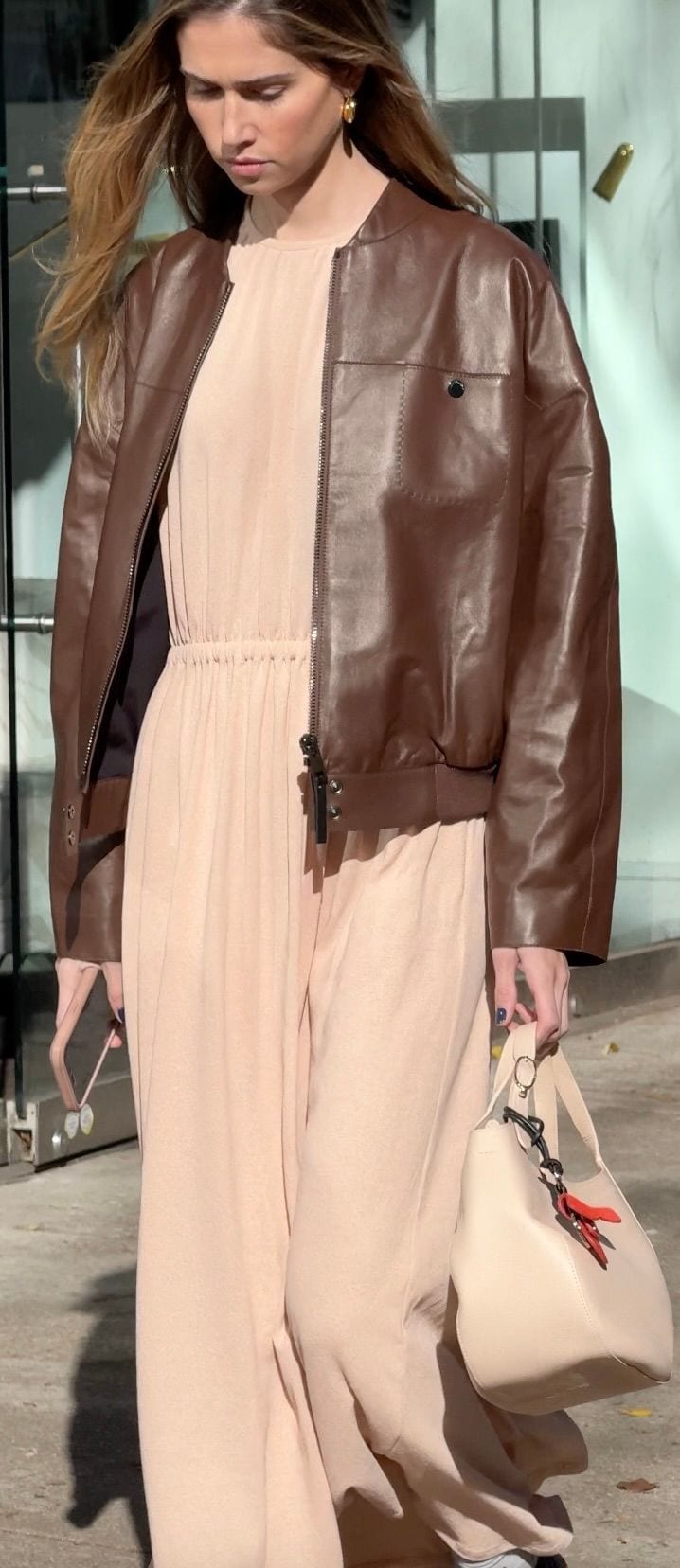 Brown leather bomber jacket over a beige jumpsuit, paired with a cream handbag.