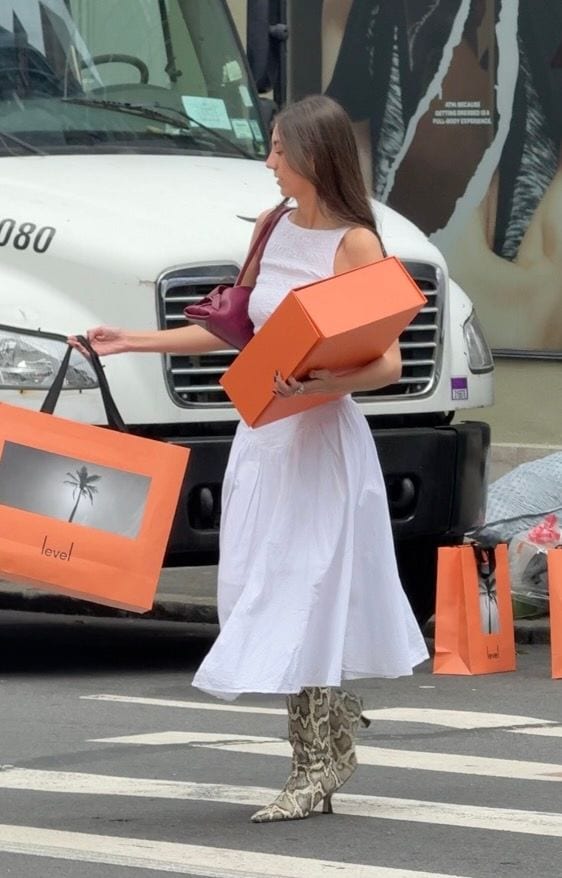 White sleeveless dress, burgundy shoulder bag, snakeskin boots, holding shopping bags and orange box.