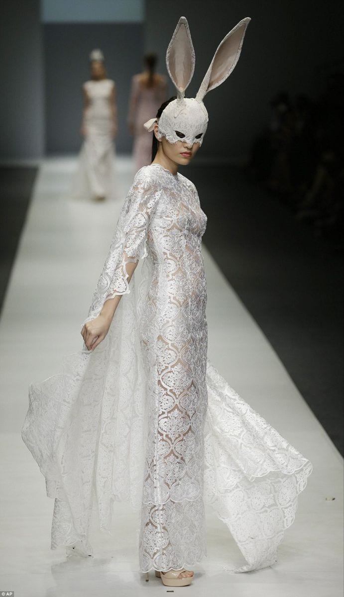 White sheer lace gown with long sleeves, bunny mask, and platform heels on runway.