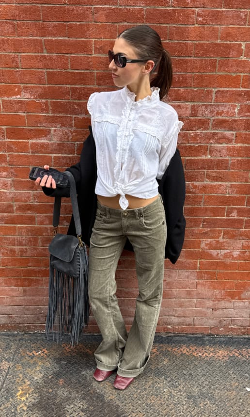 White ruffled blouse with olive corduroy pants, black blazer, fringed bag, and sunglasses.