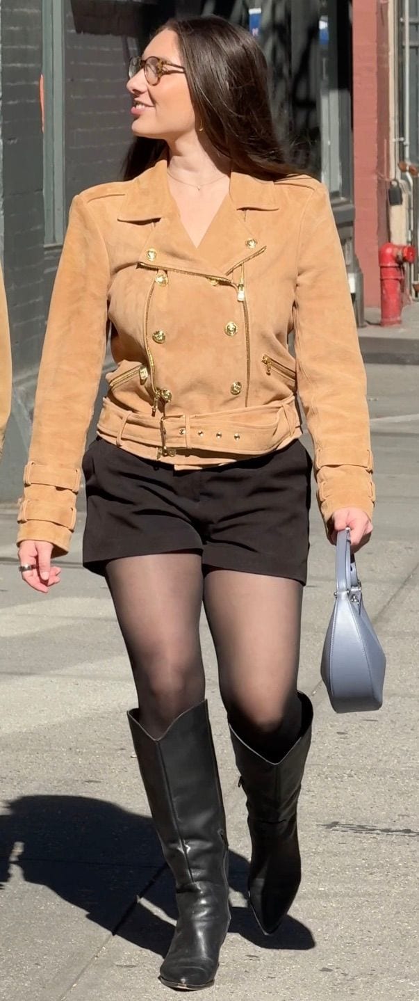 Tan suede biker jacket with gold buttons, black shorts, sheer tights, knee-high boots, and blue purse.
