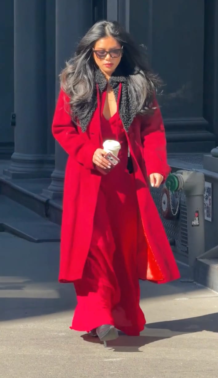 Red long coat with black faux fur collar over a red dress, carrying coffee and phone.