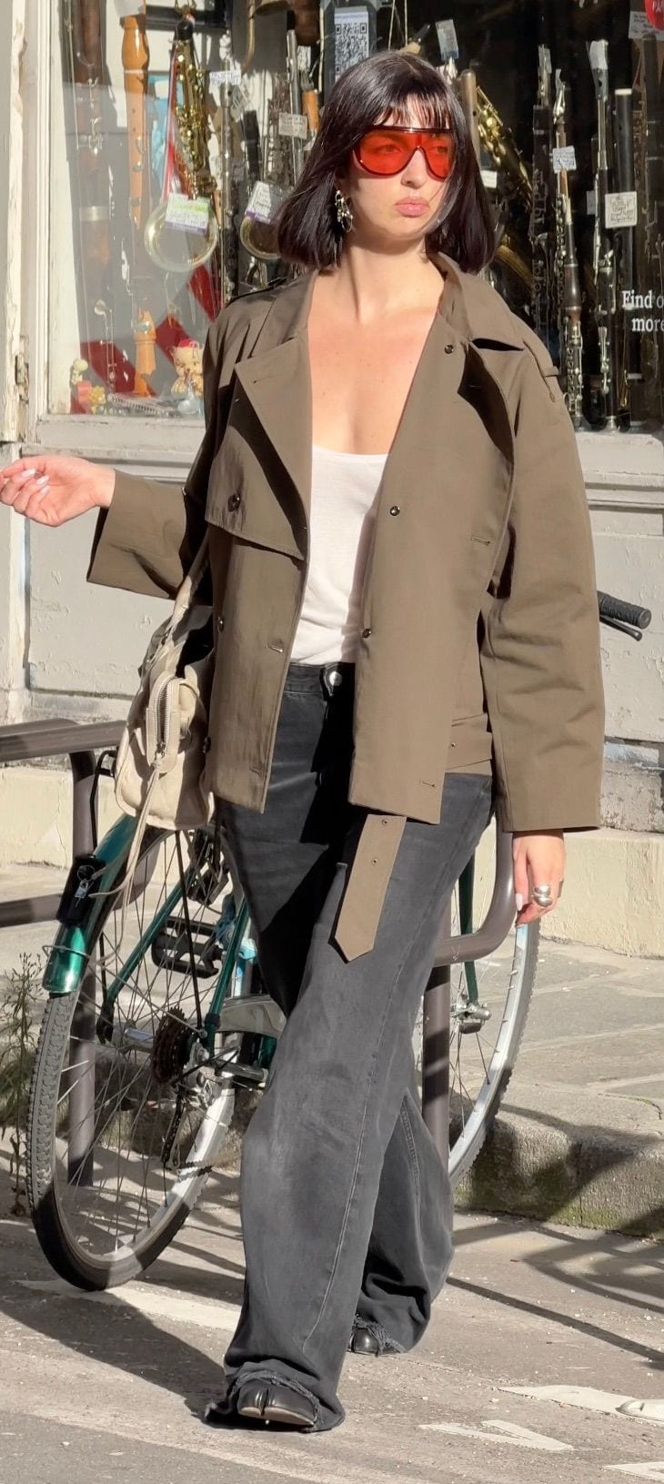 Olive green trench jacket, white tank, wide-leg black jeans, and red shield sunglasses.