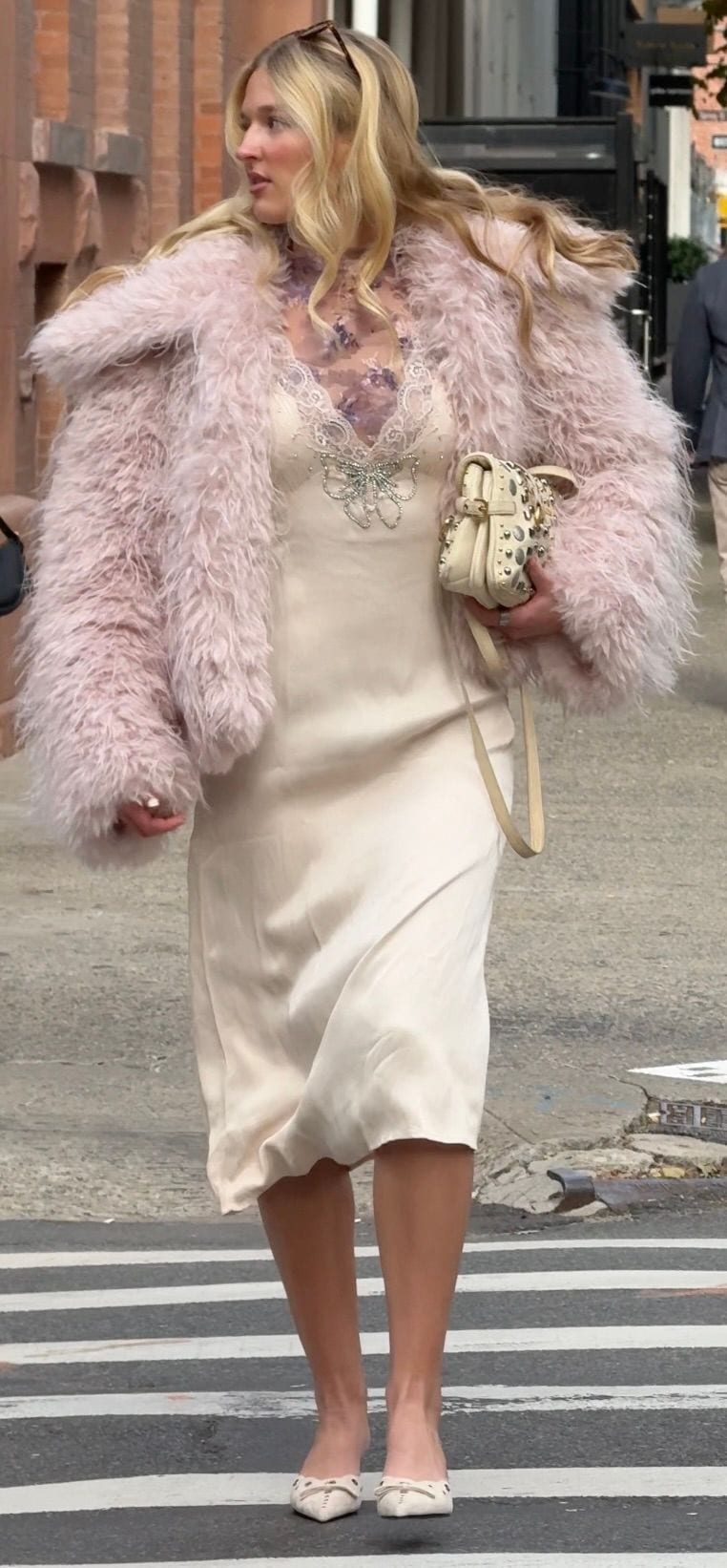 Blonde woman in a pink fluffy coat, cream slip dress with embellished butterfly, and studded bag.