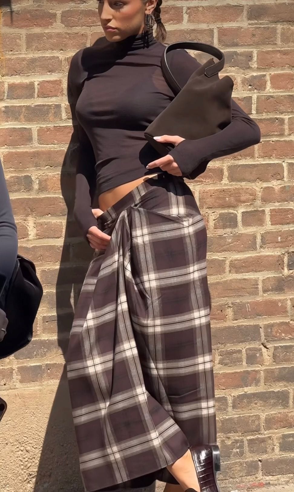 Black sheer turtleneck with brown plaid pants, brown purse, and braided hair by brick wall.