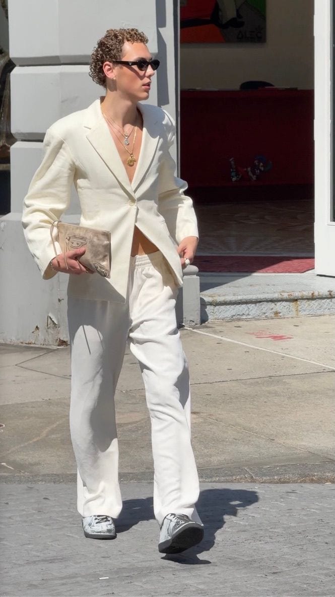 Cream suit jacket and matching trousers, holding a beige clutch, with sneakers and sunglasses.