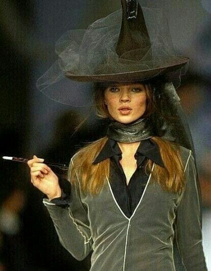 Black witch hat with veil, sheer grey coat, and black scarf, holding a cigarette holder.
