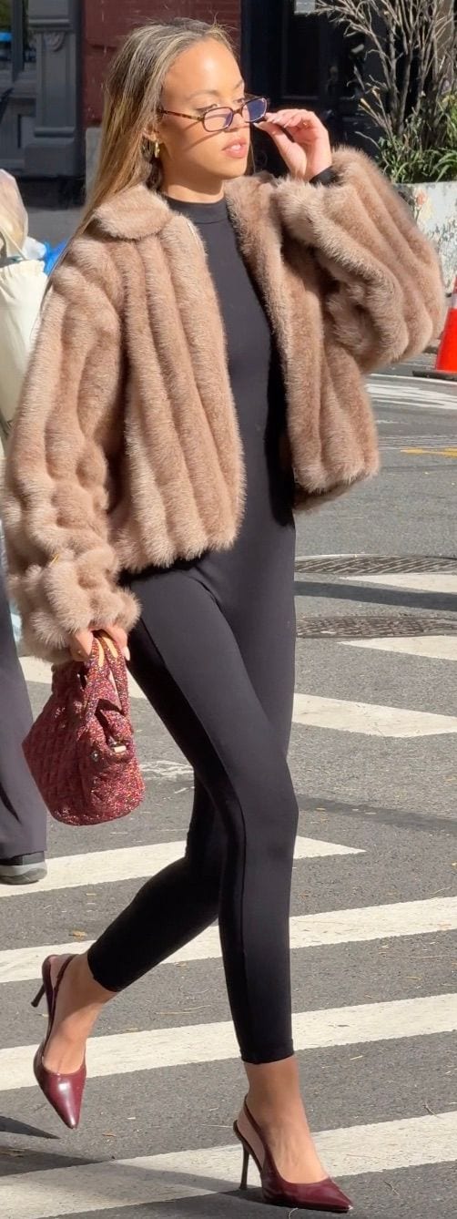 Tan faux fur jacket, black jumpsuit, red slingback heels, and a woven bag.