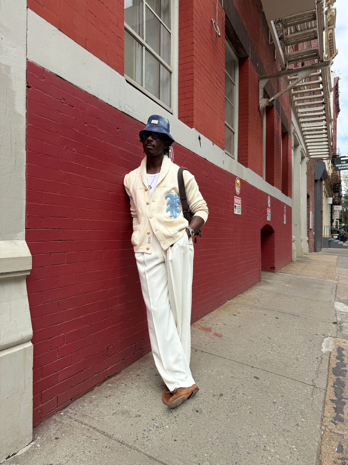 White ribbed cardigan with blue patch, wide-leg trousers, blue bucket hat, and brown shoes.
