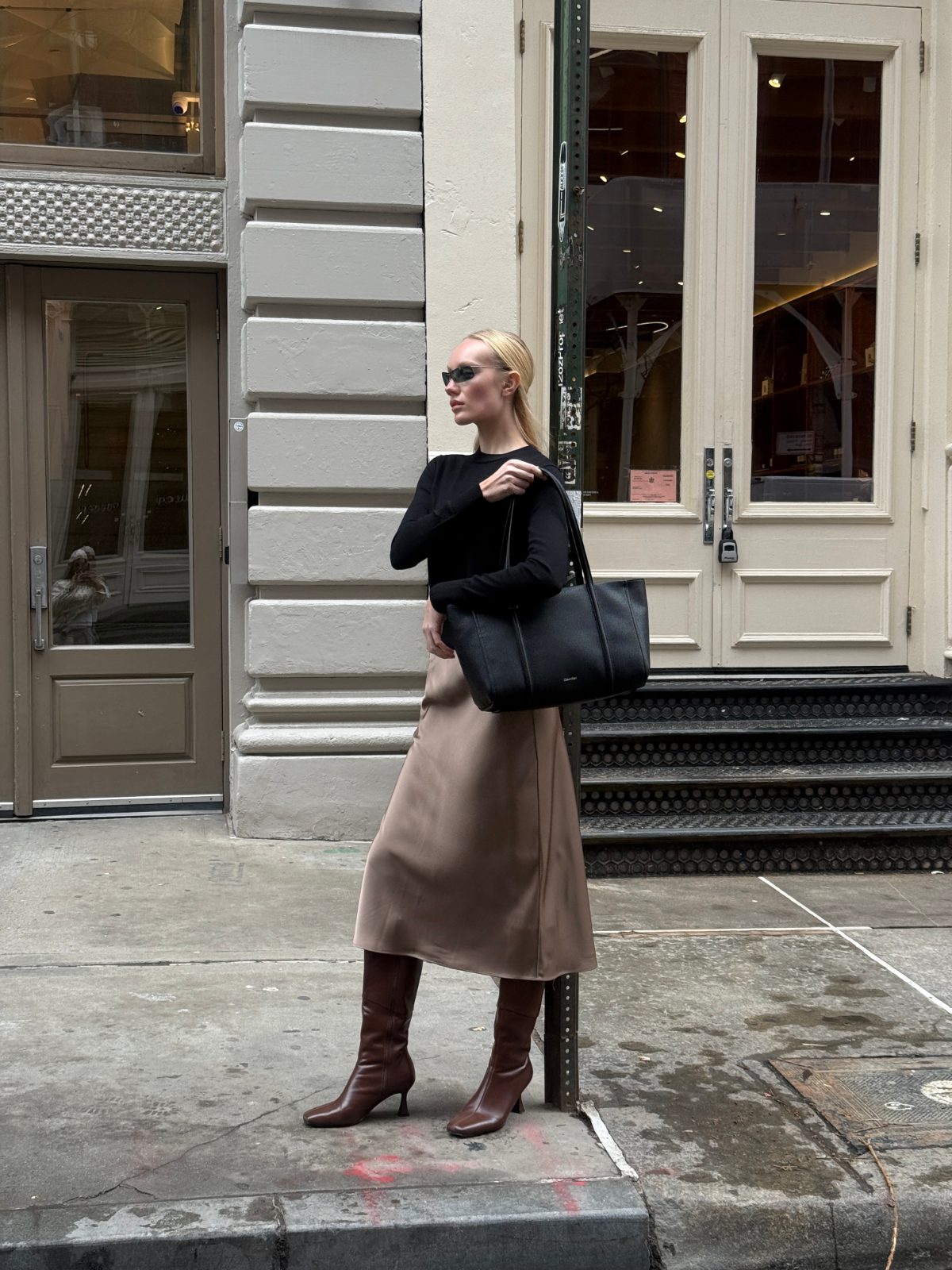 Black top, satin midi skirt, brown knee-high boots, black tote bag on a city street.
