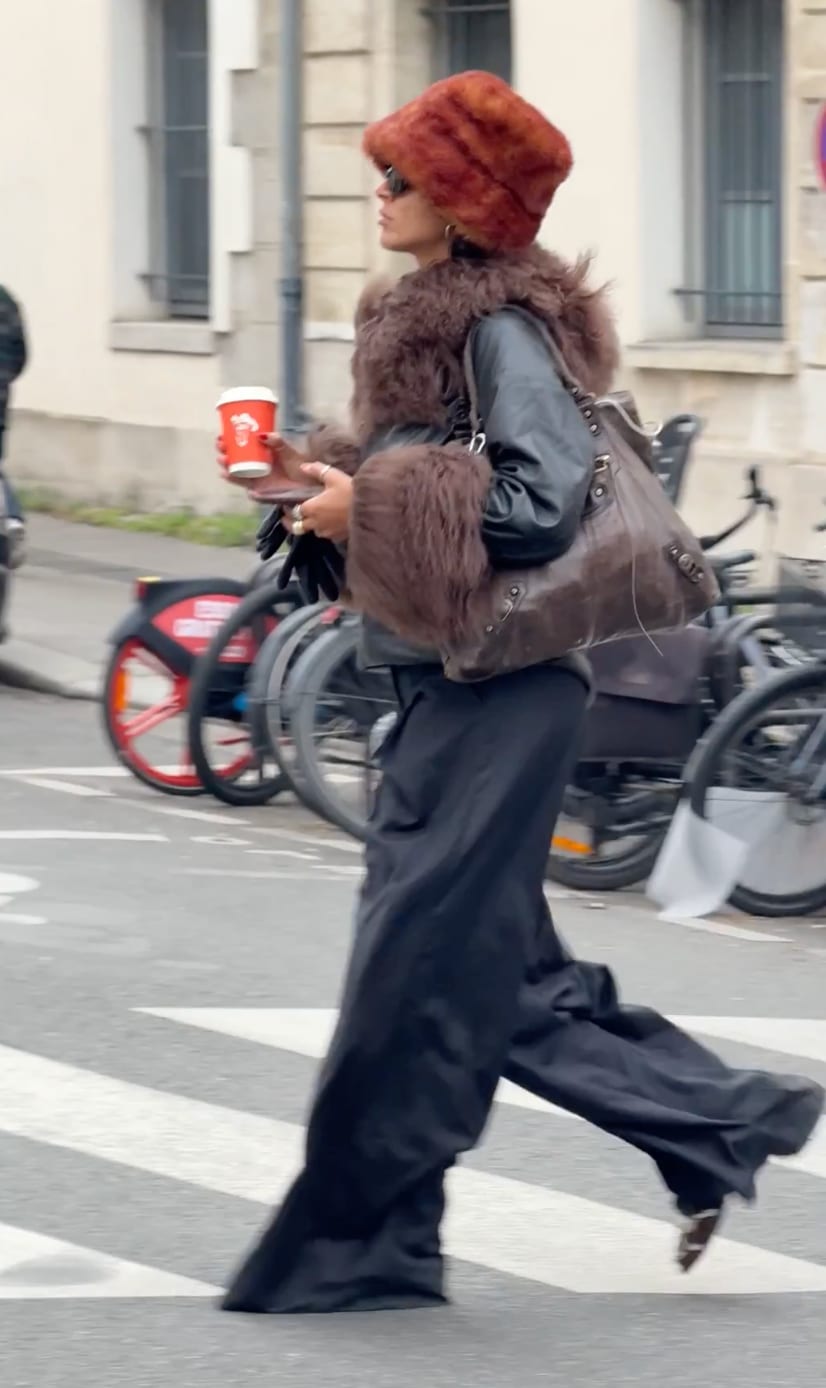 Brown fur hat, black leather jacket with fur cuffs, wide-leg pants, and a large brown bag.