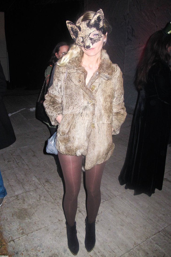 Blonde woman in fox mask, light brown fur coat, brown tights, and black ankle boots.