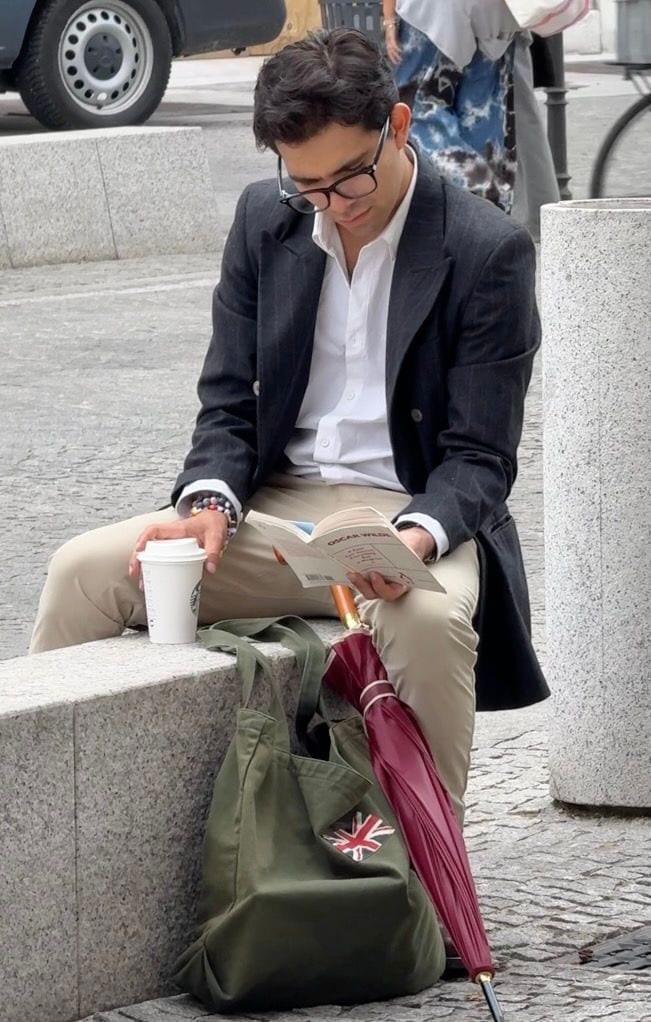 Man in dark pinstripe blazer, white shirt, and khaki pants reading book outdoors.