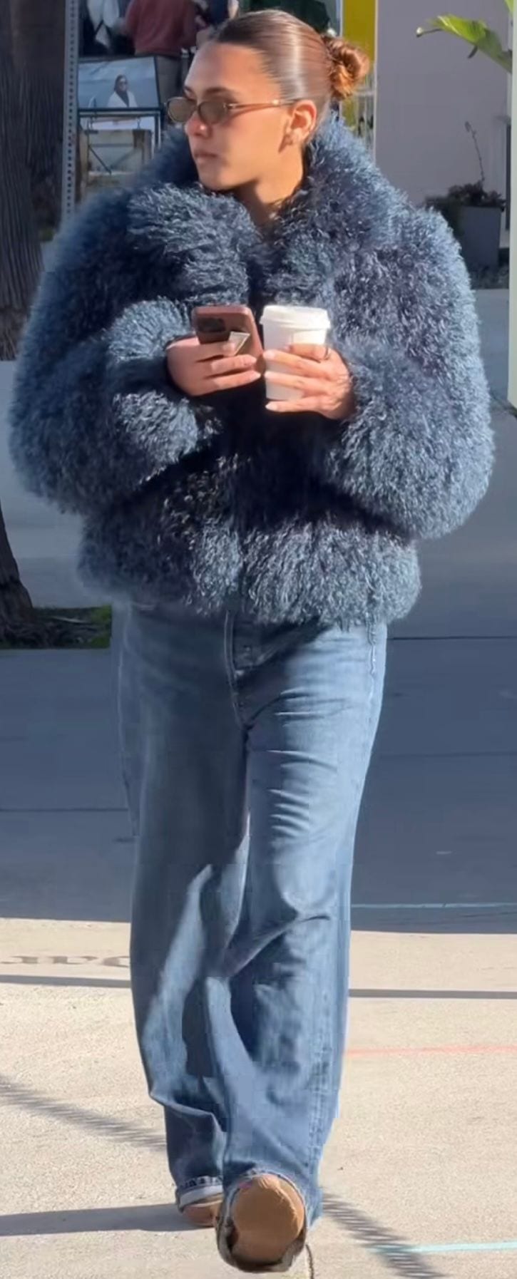 Blue fluffy coat with baggy jeans, brown sunglasses, phone, and coffee cup.