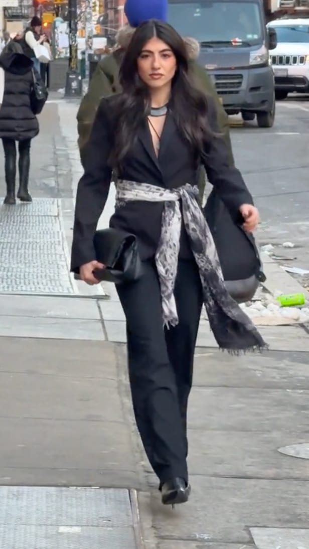 Black blazer and trousers with a gray patterned waist scarf and clutch, walking on a city street.