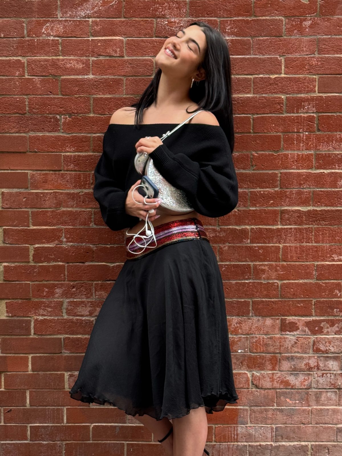 Black off-shoulder crop top, long skirt, and sequined belt against a brick wall.