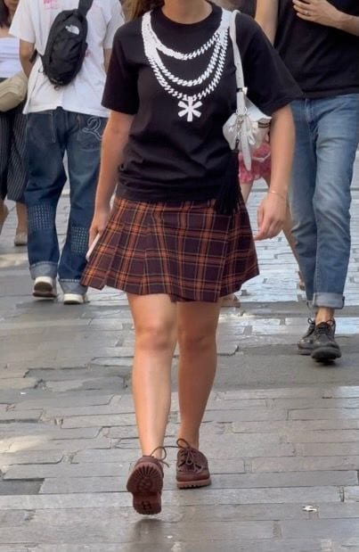 Black graphic tee with layered chain design, brown plaid mini skirt, and brown lace-up shoes.