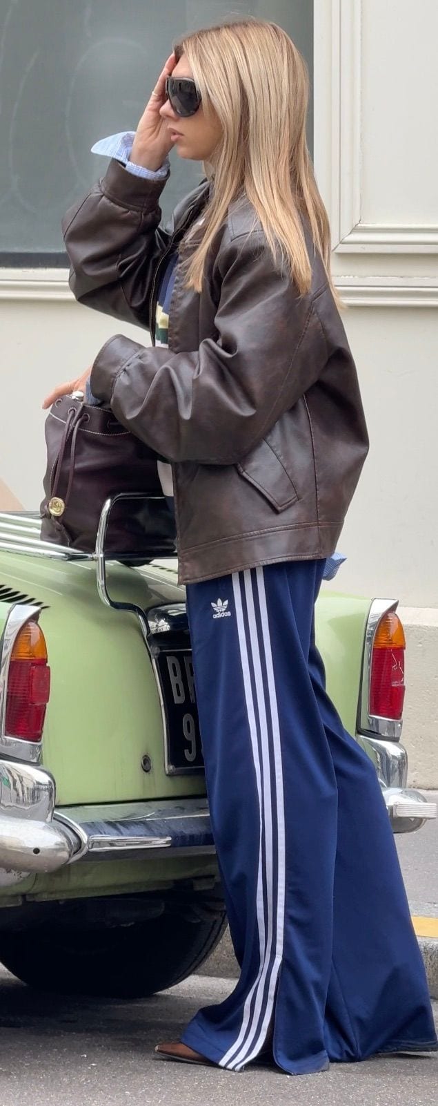 Brown leather jacket, blue Adidas track pants, and sunglasses against a light green vintage car.