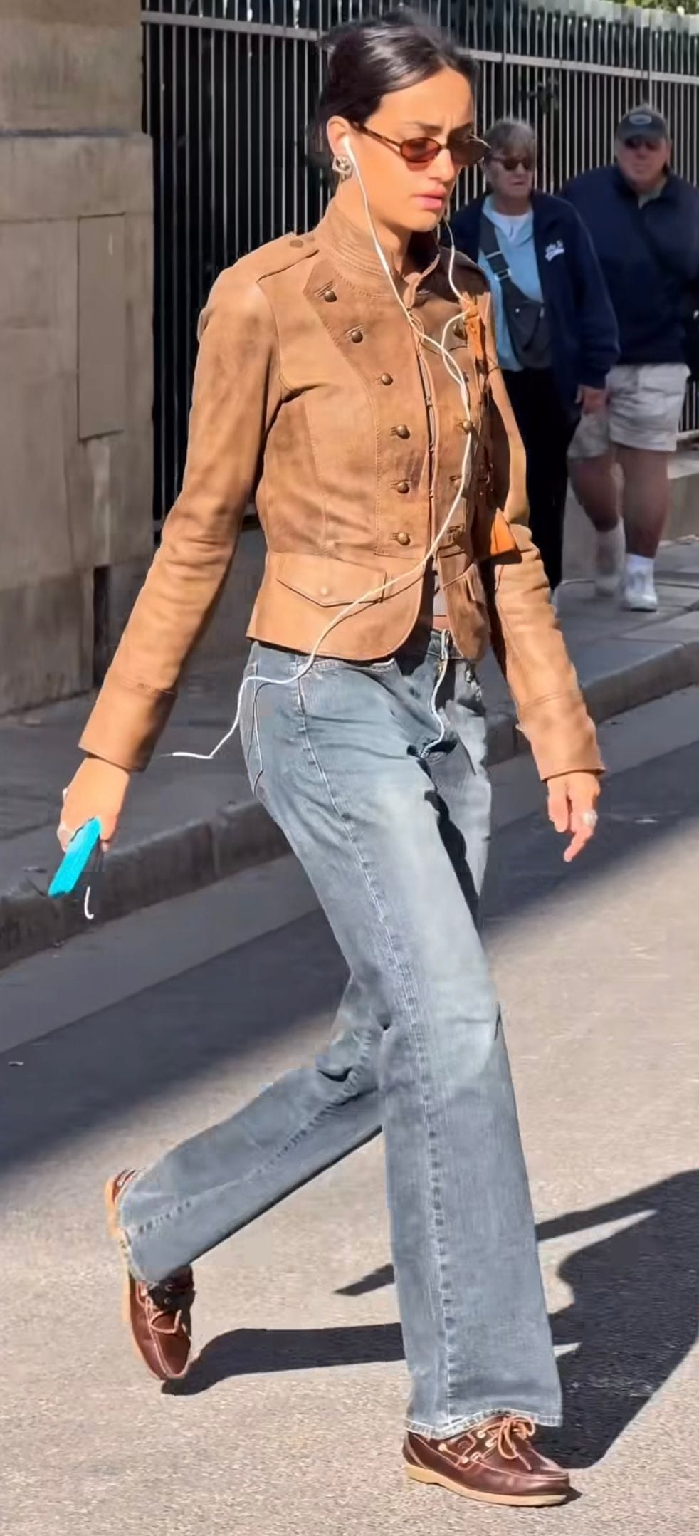 Brown military-style jacket, loose blue jeans, and brown boat shoes, with sunglasses and wired earbuds.